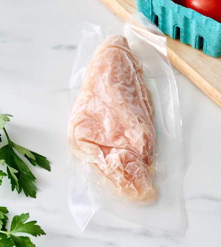 Frozen chicken breast in vacuum-sealed packaging, with fresh parsley and a basket of tomatoes in the background.